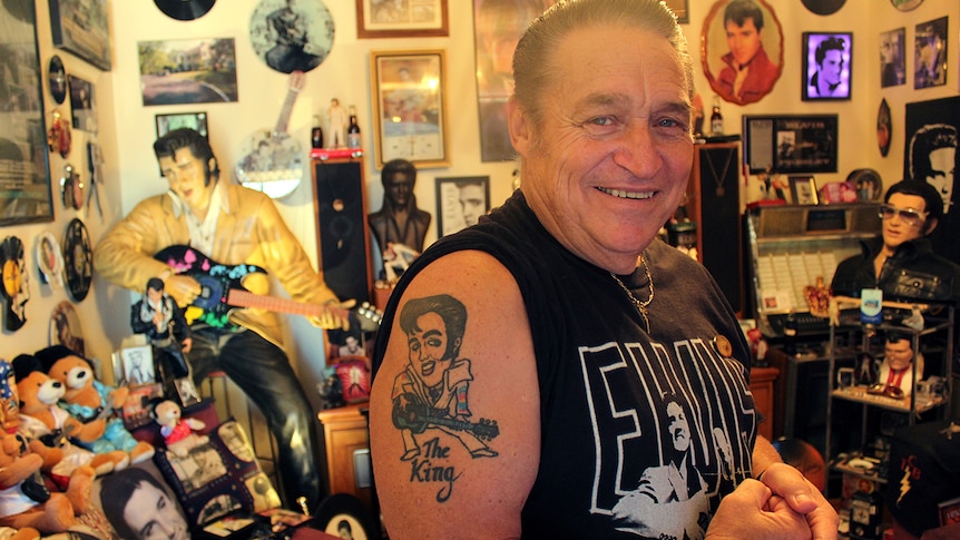 A man with slicked back hair showing off an arm tattoo of Elvis Presley surrounded by Elvis memorabilia.