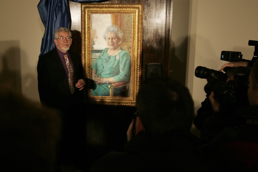 A white-haired man with dark glasses stands next to a portrait of Queen Elizabeth II as photographers take pictures.