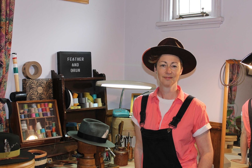 Woman with short blonde hair wearing brown hat smiles next in her hat making studio.