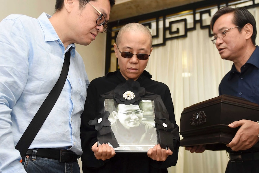 Liu Xia, the wife of Liu Xiaobo, stands between two men while holding a portrait of her late husband.