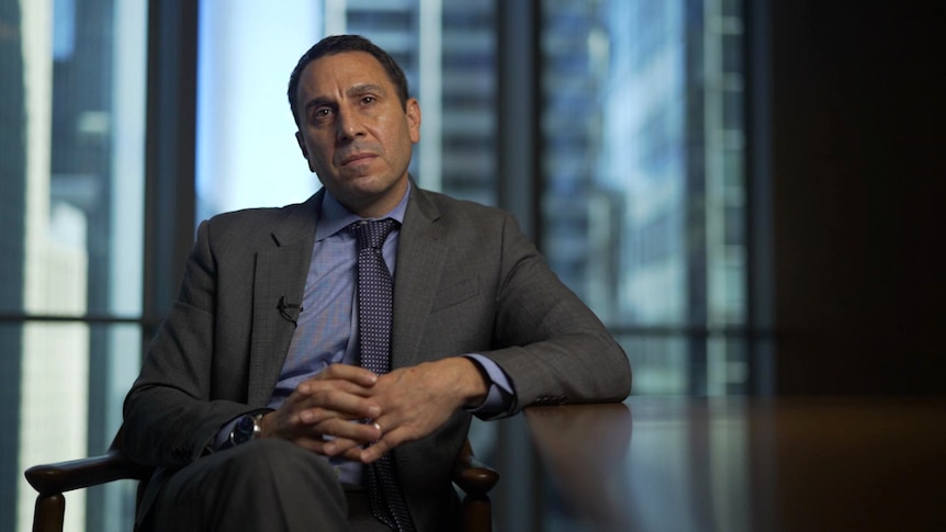 A man wearing a suit leans against a boardroom-style table in an office, city buildings can be seen through the window.