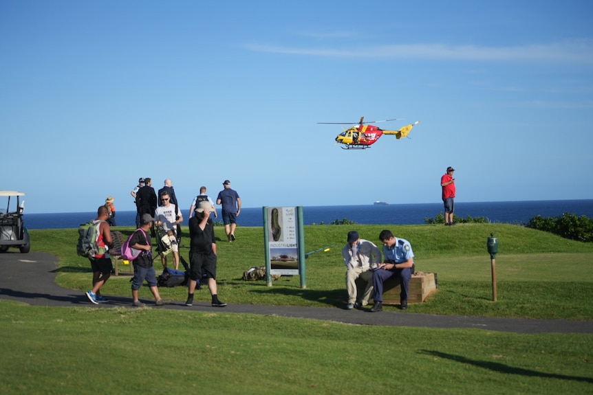 A police officers talks to a man on a headland while a helicopter hovers in the air behind. 