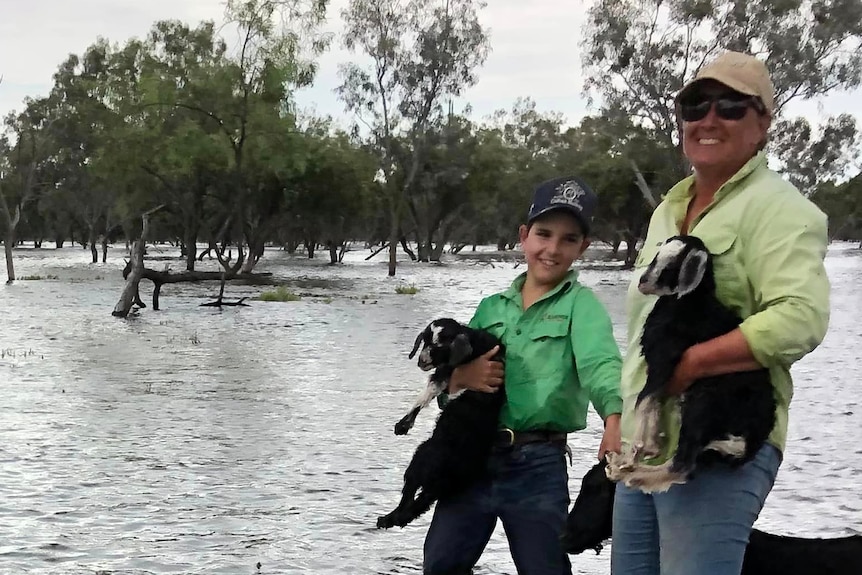 A woman and a boy standing in a flooded paddock holding some goats