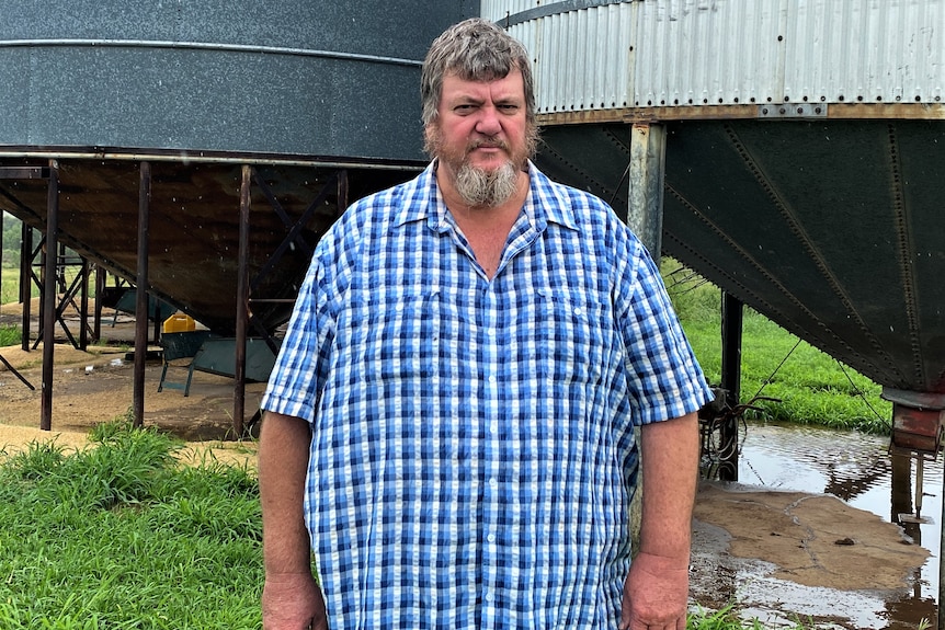 A man with grey hair and a long grey beard looks concerned. He's standing in the rain in front of grain silos  
