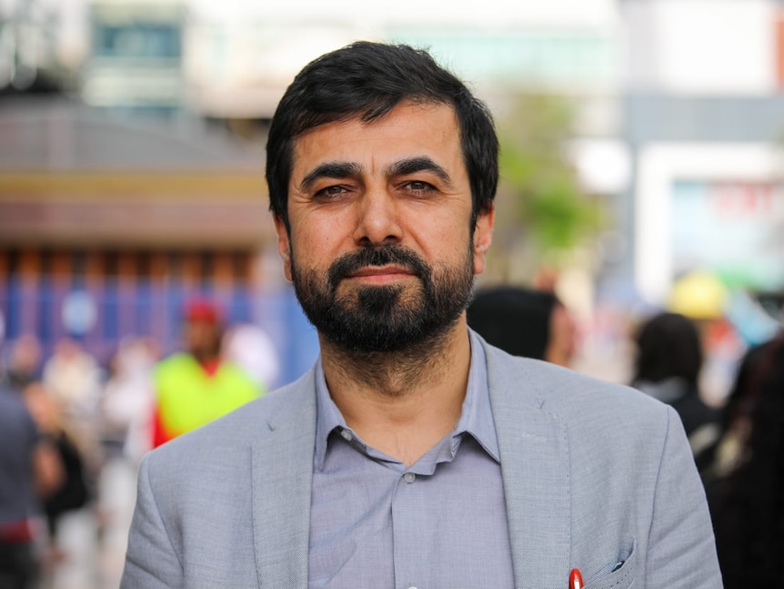 A photo of a man wearing a light-coloured jacket and shirt with people in the background.