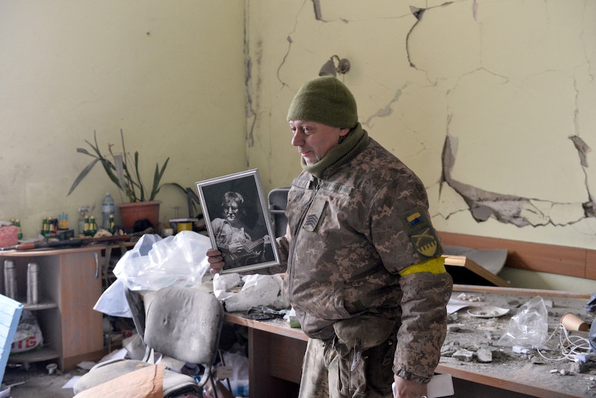 In a damaged room a man in military fatigues holds up a small black and white portrait of John lennon