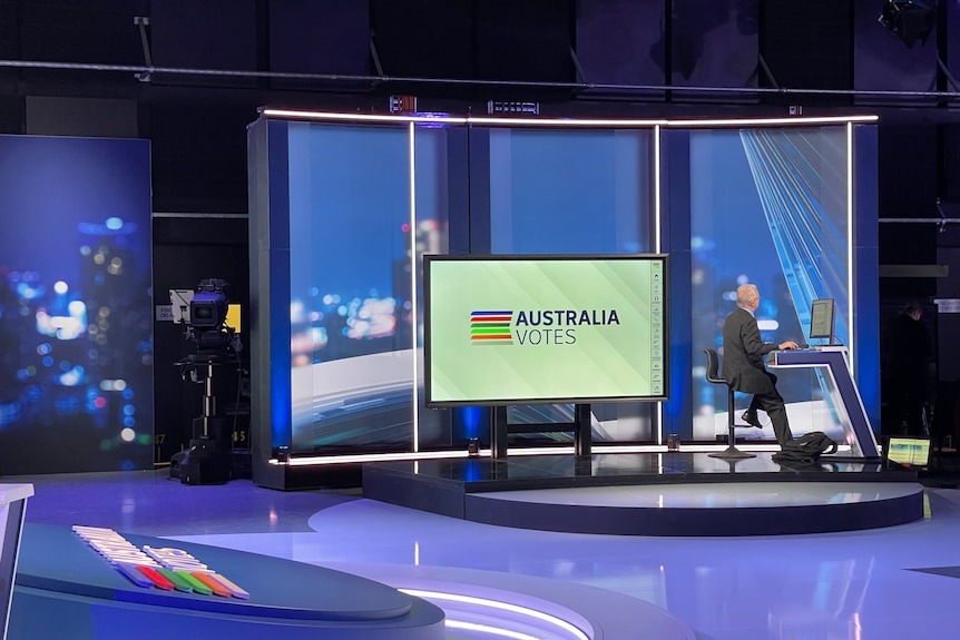 Wide shot of a TV studio with a man sitting at a laptop.