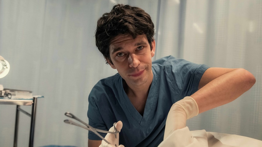 A TV still of Ben Whishaw, looking into the camera. He's wearing blue scrubs and surgical gloves and is holding forceps.