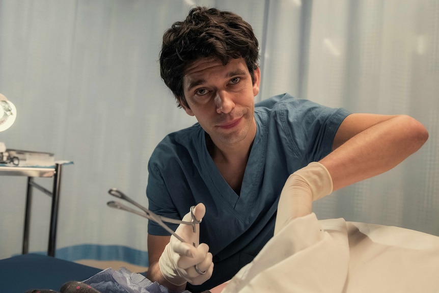 A TV still of Ben Whishaw, looking into the camera. He's wearing blue scrubs and surgical gloves and is holding forceps.