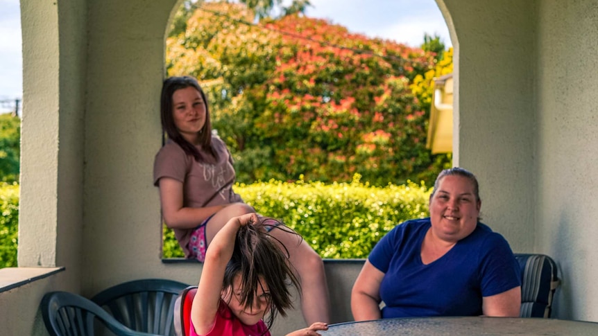 A woman sits on chair on a verandah smiling with a teenage girl and a young girl pulling a face next to her.