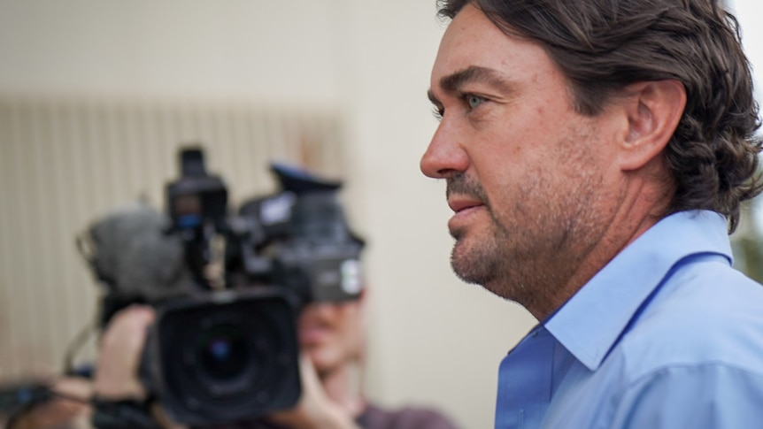 Side-profile mid-close-up of a white man in a light blue collared shirt, walking past a big news camera.