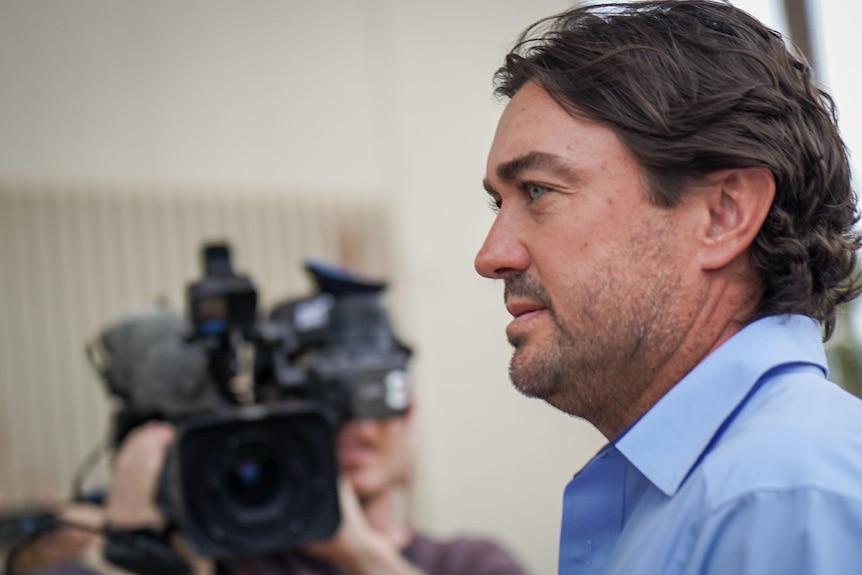 Side-profile mid-close-up of a white man in a light blue collared shirt, walking past a big news camera.