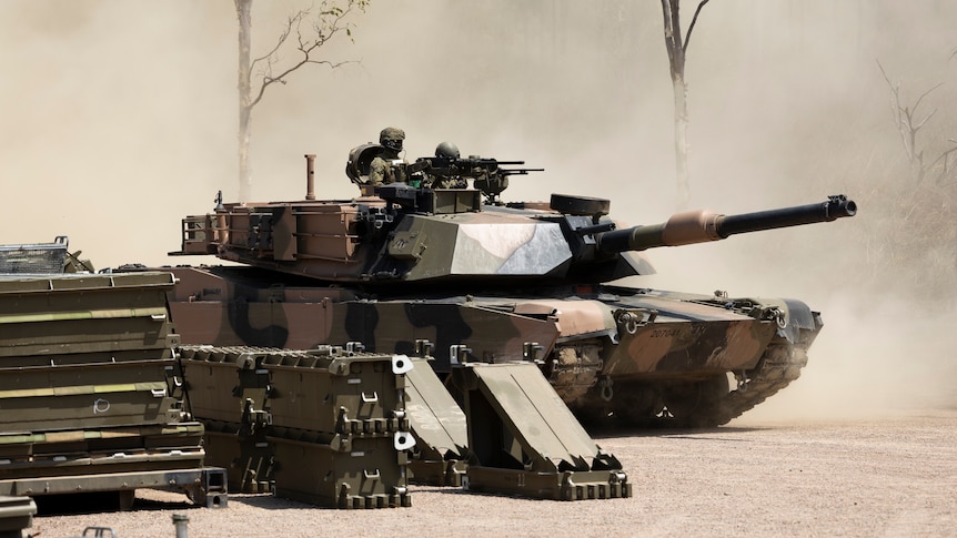 A military tank in camouflage print. There's a man in military uniform standing on it.