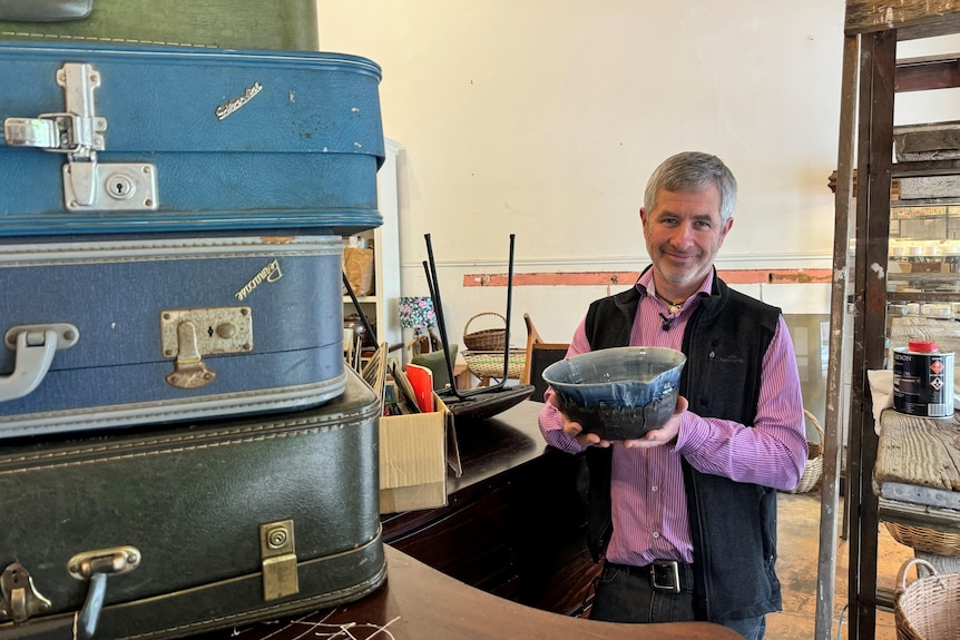 A man with a greying beard holds a blue-coloured bowl behind a table with drum cases inside a store.
