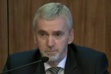 Ken Jones wears a suit with a grey and black striped tie as he sits in front of a microphone giving evidence to the commission.