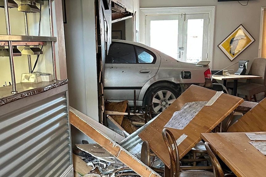 The back of a silver car lodged in a wall in the pub. it is morning, and sun is shining on the dining tables next to the kitchen