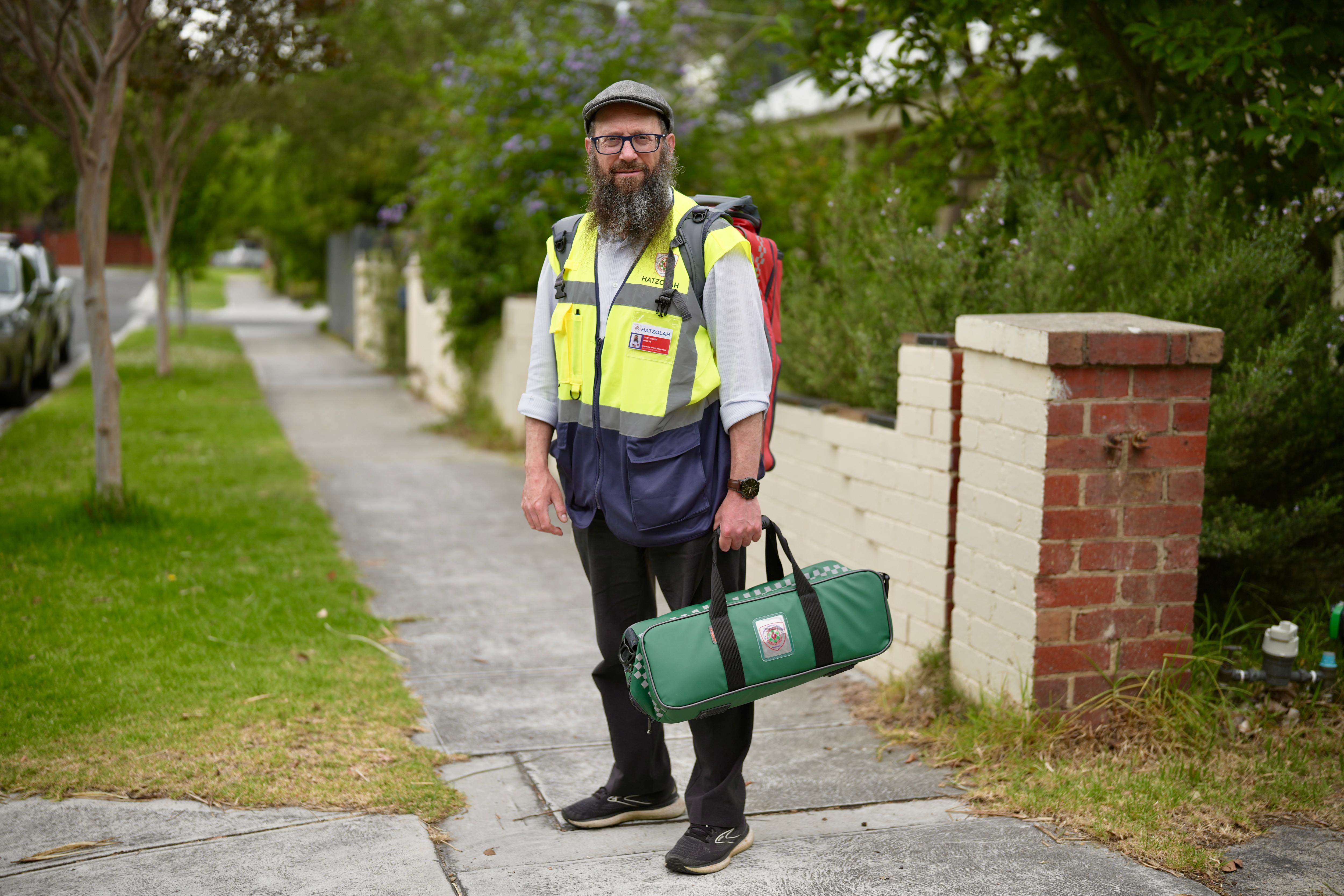 Um homem de barba longa e óculos, boné cinza e colete amarelo de alta visibilidade está em uma trilha segurando uma bolsa verde.
