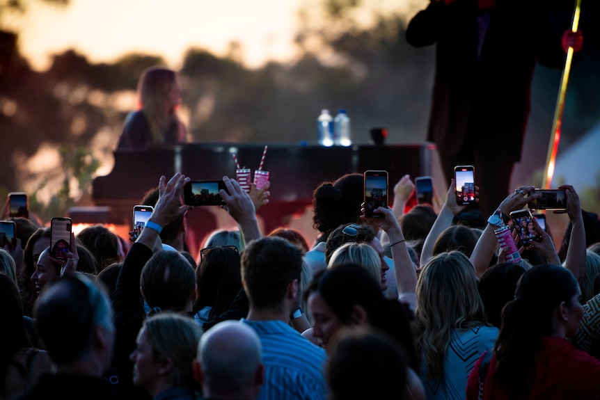 A crowd of people with some holding up their phones face a stage with a person playing piano