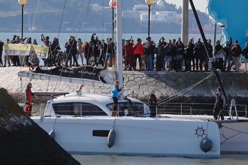 A boat sails into a harbour with crowds of people gathered around.