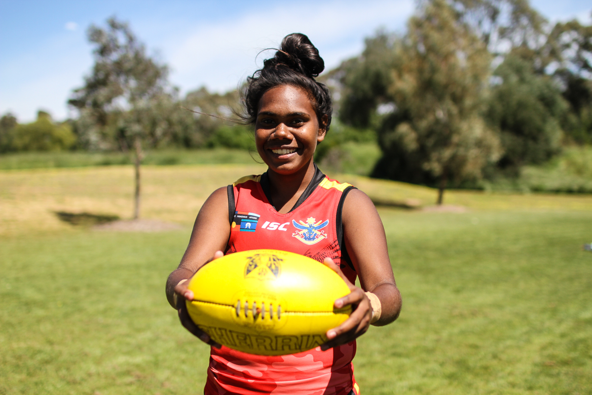 Julianna Kerinaiua, 17 from the Tiwi Bombers Football team stands smiling and holding a football.
