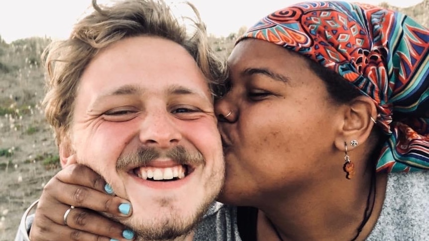 Man smiles at the camera while his girlfriend kisses him on the cheek, to depict talking about race in interracial relationships