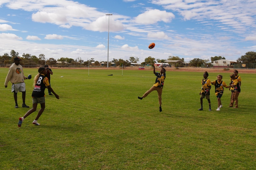 An Indigenous boy jumps in the air to catch an AFL ball, while other children and people watch.