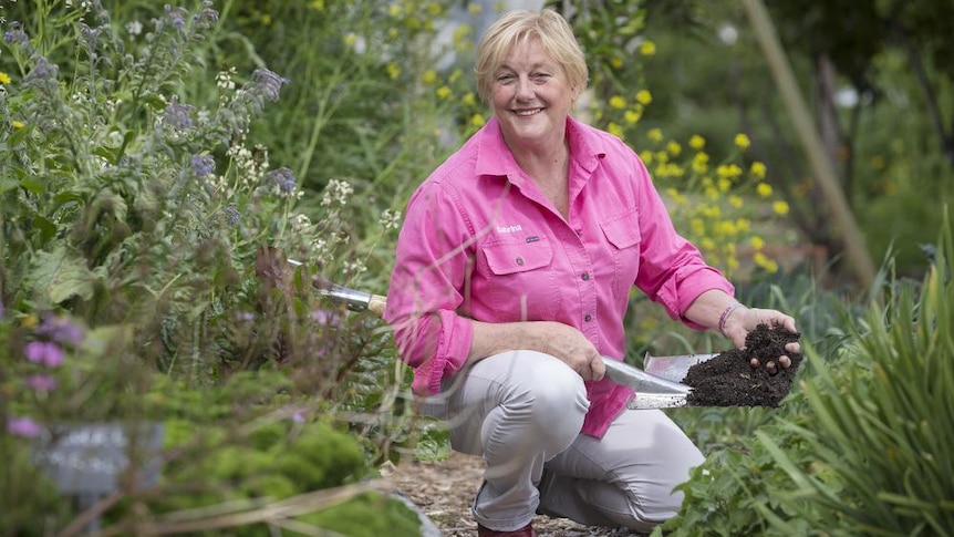 A woman in gardening clothes crouched in a garden holding soil.