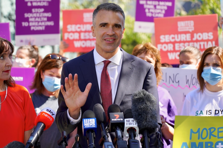 A man stands in front of microphones with his hand up. Behind him are people wearing masks and holding signs