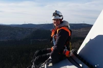 A woman in a white hard hat and work clothes sits on a white wind turbine blade above a forest