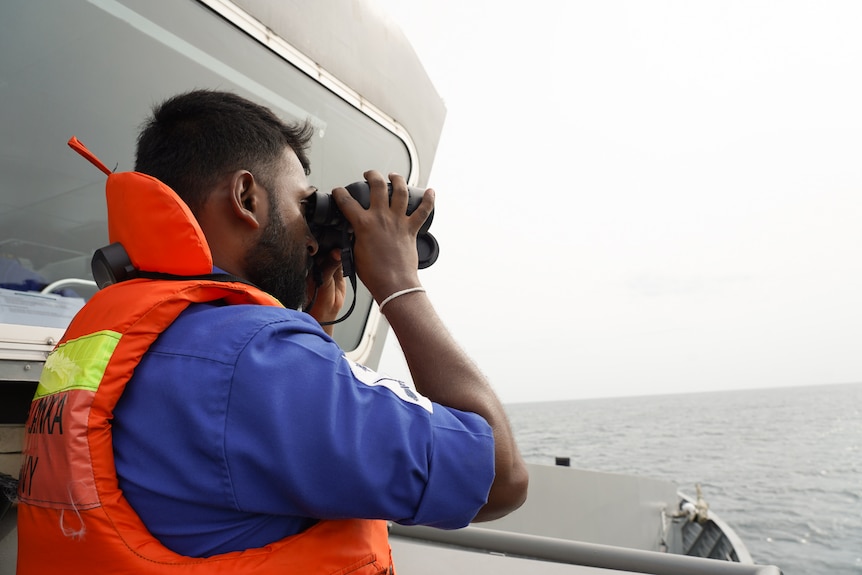 A man wearing a life vest looks through binoculars over the ocean.