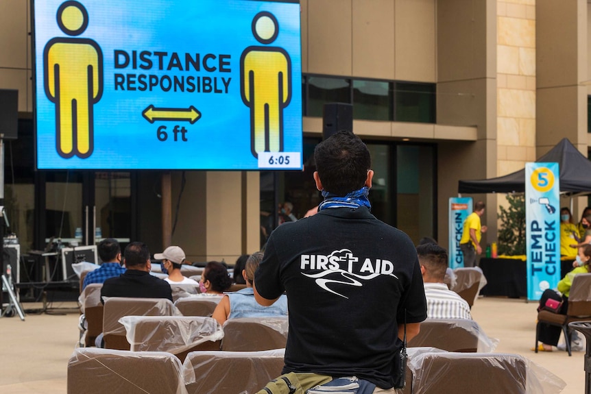 People sit on plastic covered chairs outside a building with a big screen displaying a coronavirus safety message.