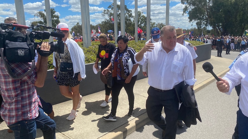 Craig Kelly speaking to news crews while walking with three protesters away from the large group 