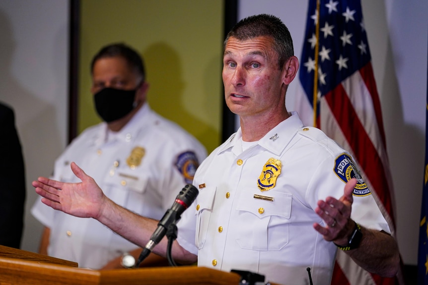 craig mccartt in police uniform holds his arms out wide as he speaks at a lectern with a fellow officer and a flag behind him