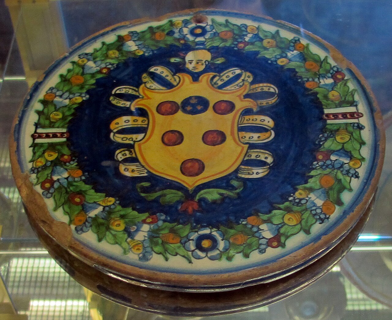 A photo of a circular wall plaque showing a coat-of-arms featuring 5 red balls and a blue ball on a golden shield
