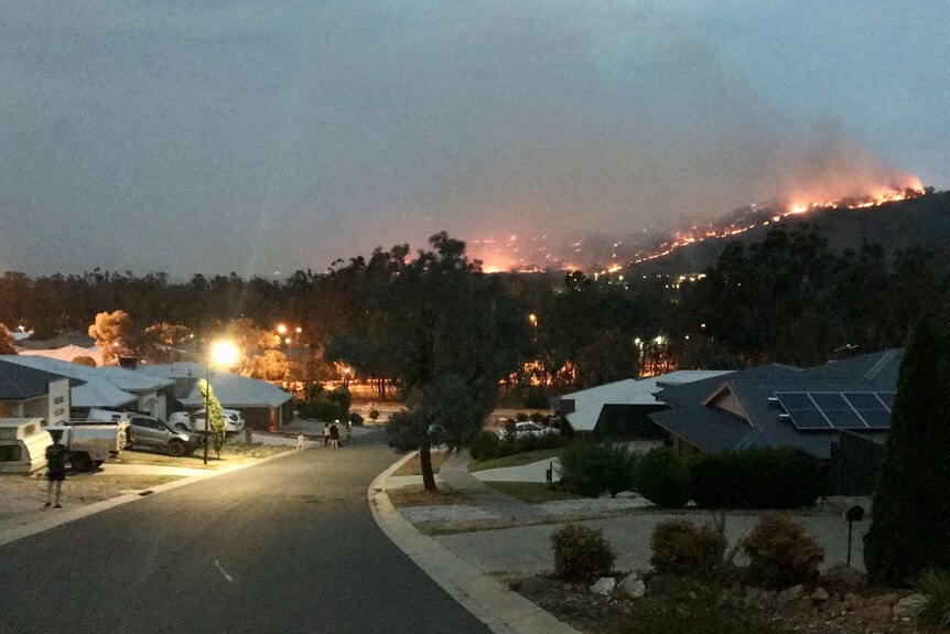 A fire burns on a hill near a suburban street in the evening.