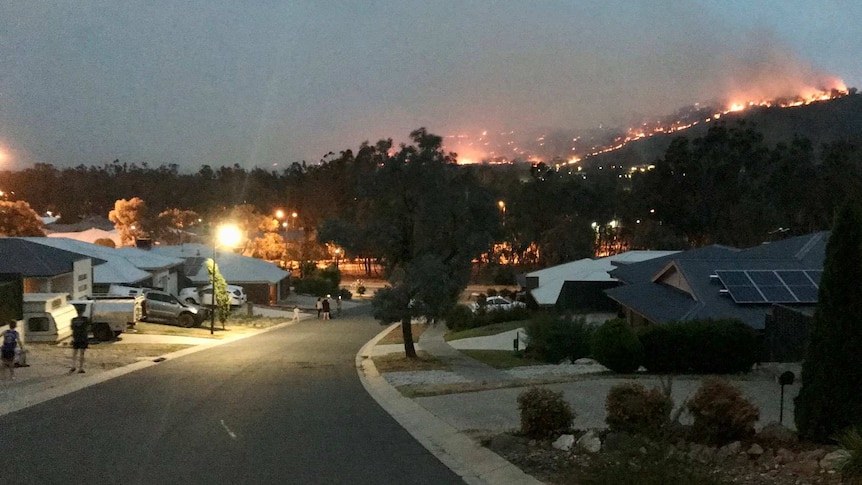 A fire burns on a hill near a suburban street in the evening.