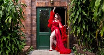 A woman strikes a pose in a doorway. She wears a red corset and briefs covered with a red translucent gown with furry cuffs.