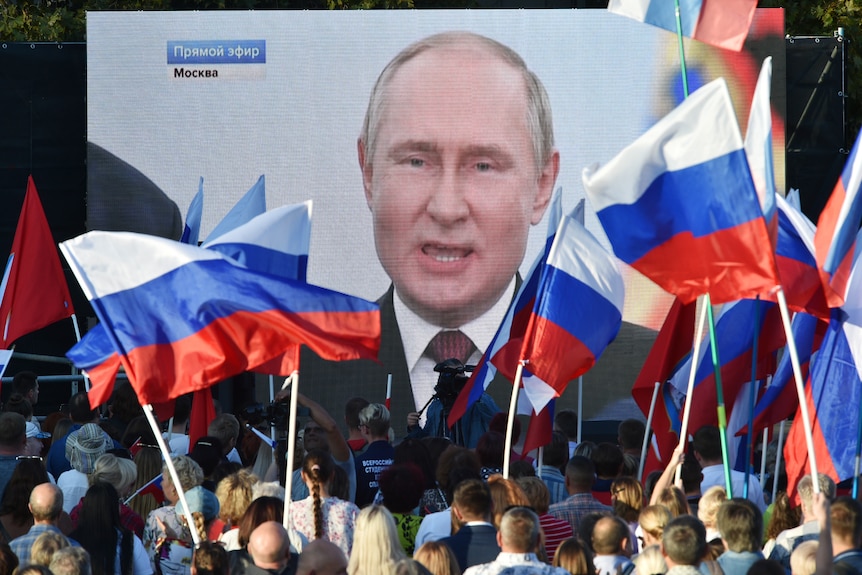 Vladimir Putin speaking on a large screen to a crowd waving large Russian flags