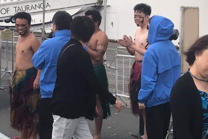 A group of shirtless men clap their hands at a ship port. They wear grass skirts and have black lines painted on their faces.