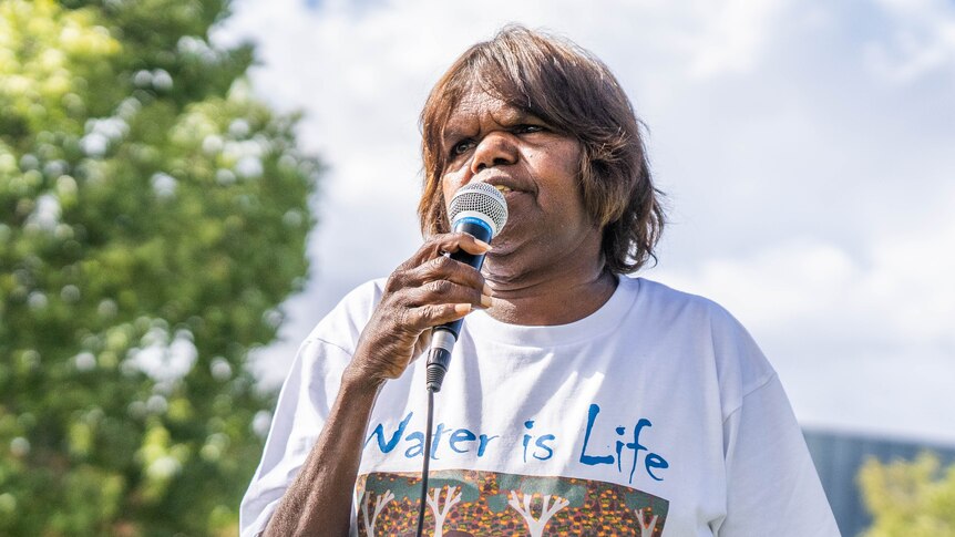 A woman speaks into a microphone. Her t-shirt reads 'water is life'.