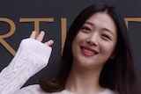 An asian woman in a white top smiles and waves for the camera.