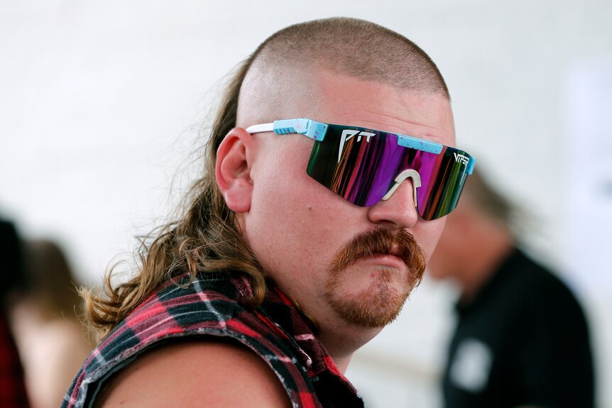 A man with a mullet and coloured sunglasses looks at the camera.