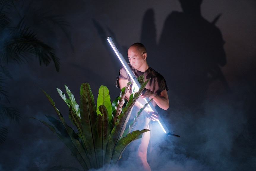 A bald Asian man holds a light bar in his hands as he dances on stage. He is holding the light to a plant.