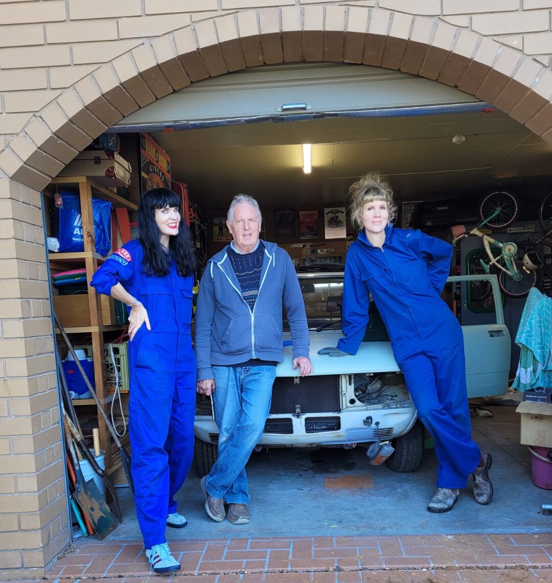 Three people stand, two in blue overalls, in front of Fiat 132 restoration project