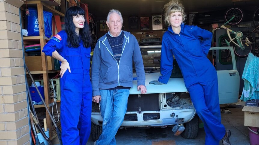 Three people stand, two in blue overalls, in front of Fiat 132 restoration project