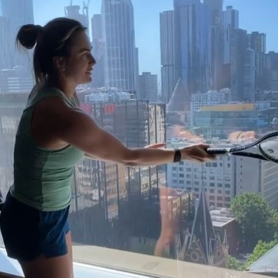 A woman in a singlet and shorts hits a tennis ball against a window.