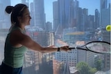 A woman in a singlet and shorts hits a tennis ball against a window.