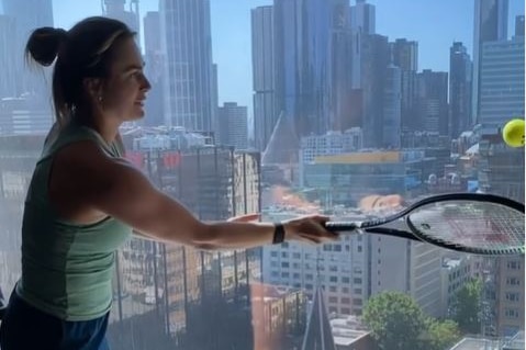 A woman in a singlet and shorts hits a tennis ball against a window.