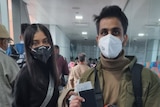 An Indian woman and a man standing waiting to get on a plane wearing masks, the man is holding up his passport