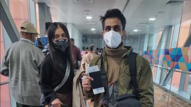 An Indian woman and a man standing waiting to get on a plane wearing masks, the man is holding up his passport